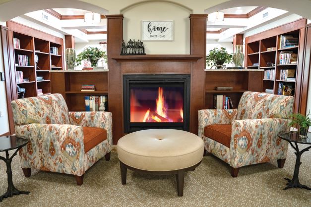 A cozy seating area featuring two patterned armchairs with orange cushions, a round beige ottoman, and a lit fireplace. The background has wooden bookshelves filled with books and decorative plants. A sign above the fireplace reads 