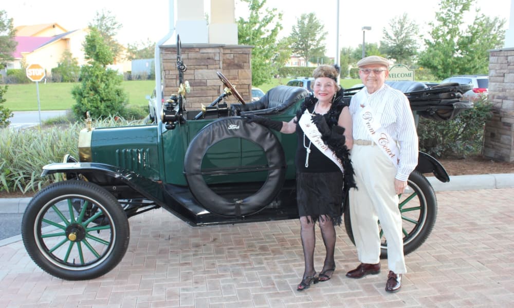 A smiling older couple dressed in vintage attire stands beside a green antique car. The woman wears a black dress and feathered headband, while the man is in a light shirt and pants with a cap. Both have sashes, and the scene is outdoors with greenery in the background.