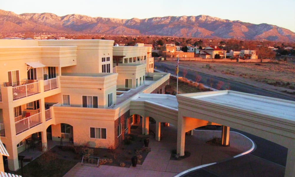 A beige, multi-story retirement community building with verandas and arches is shown at sunset, with a mountain range in the background and a curving driveway in the foreground. The sky is clear, casting a warm glow over the scene.