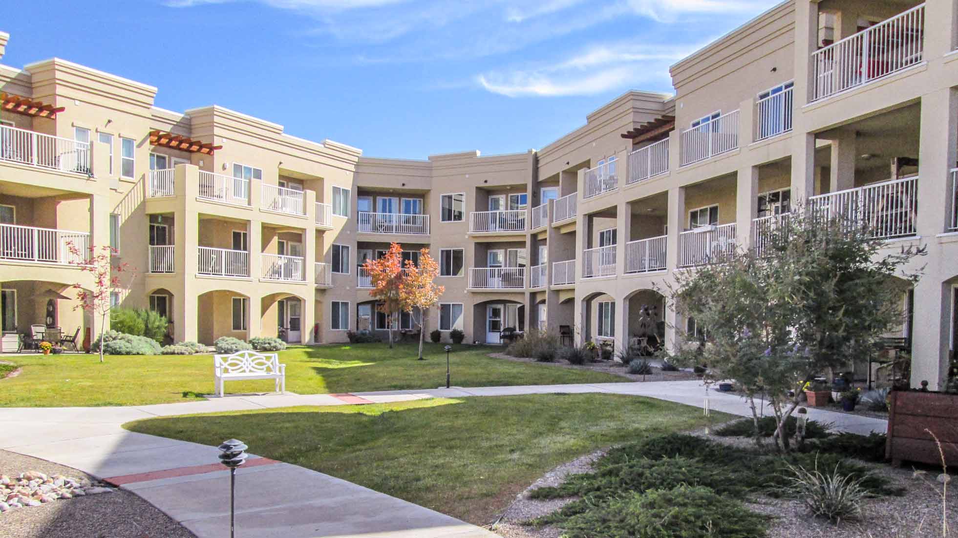 A three-story beige apartment building with balconies surrounds a landscaped courtyard featuring grass, walking paths, shrubs, a white bench, and a few small trees under a blue sky.