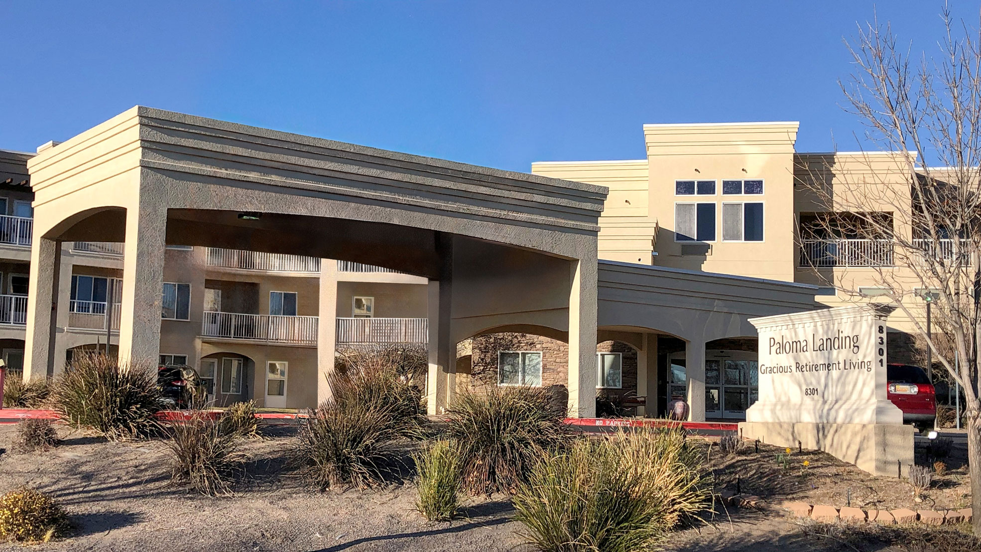 A modern retirement living facility with a large covered entrance, beige exterior, balconies, and landscaped grounds with dry grass and shrubs. A sign in front reads "Paloma Landing Gracious Retirement Living.