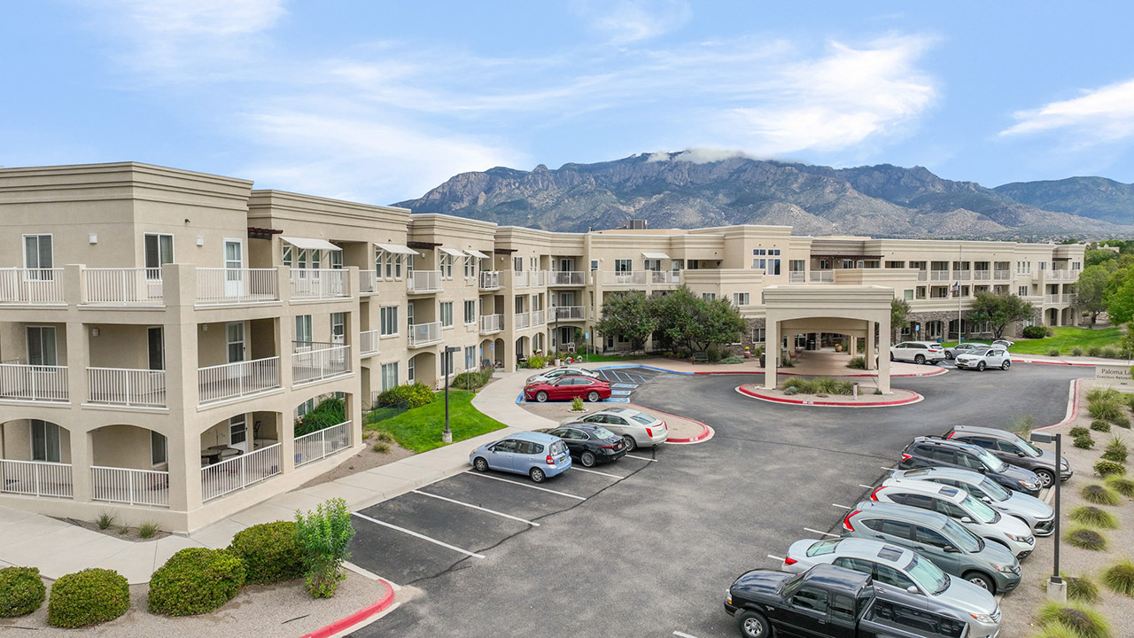A large beige three-story apartment building with balconies and arched windows, surrounded by parked cars and landscaped greenery, with mountains and a blue sky in the background.