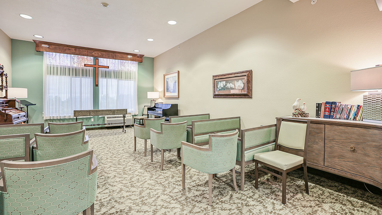 Small chapel room with green upholstered chairs arranged in rows, a piano, a wooden cross on the wall by large windows, a sideboard with books and a lamp, and soft neutral-colored walls and carpet.