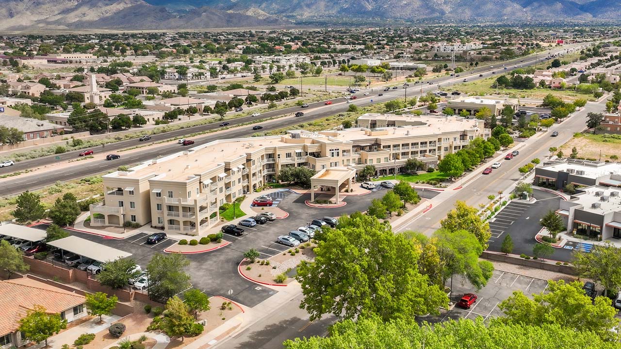 Aerial view of a large beige building, possibly an apartment complex or hotel, with a parking lot, surrounded by trees, roads, and other buildings, set against a backdrop of mountains and a suburban landscape.