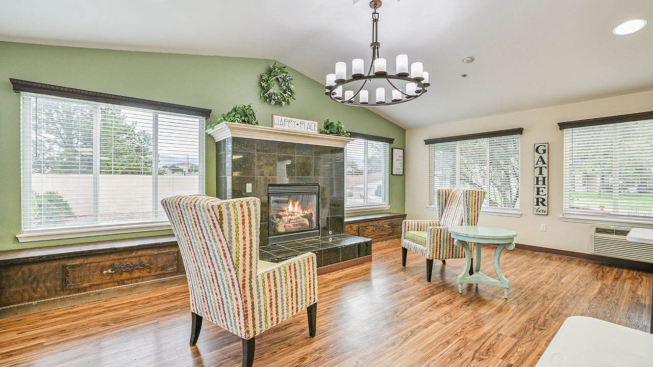 Bright living room with hardwood floors, two patterned armchairs by a black-tiled fireplace, round mint table, large windows with white blinds, and a decorative chandelier above. A “GATHER” sign hangs on the wall.