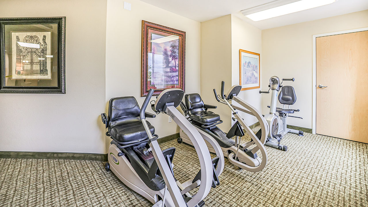 Two stationary recumbent exercise bikes in a small, carpeted fitness room with several framed pictures on the walls and a closed wooden door in the background.