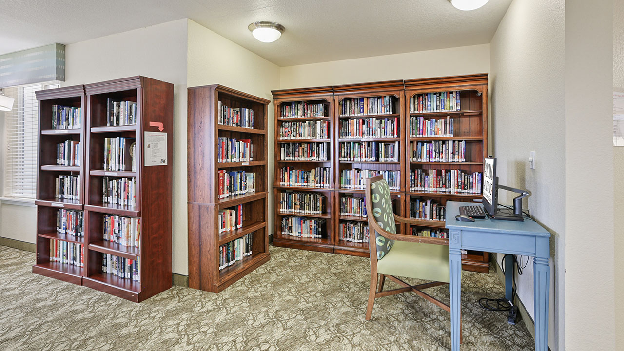 A small library room with three tall bookshelves filled with books, a desk with a computer, and a patterned chair on a light-colored carpeted floor. Bright lighting and cream-colored walls.