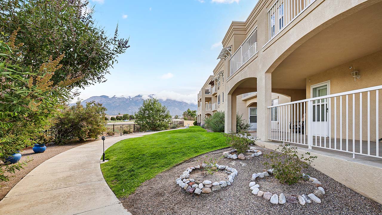 A landscaped outdoor area with a curved sidewalk, green lawn, and small rock garden beside a beige apartment building with balconies; mountains and blue sky are visible in the background.