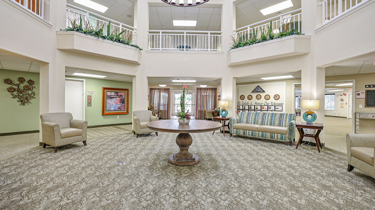 Spacious lobby with a round wooden table in the center, surrounded by armchairs, a sofa, decorative plants, artwork on the walls, and natural light coming from large windows above.