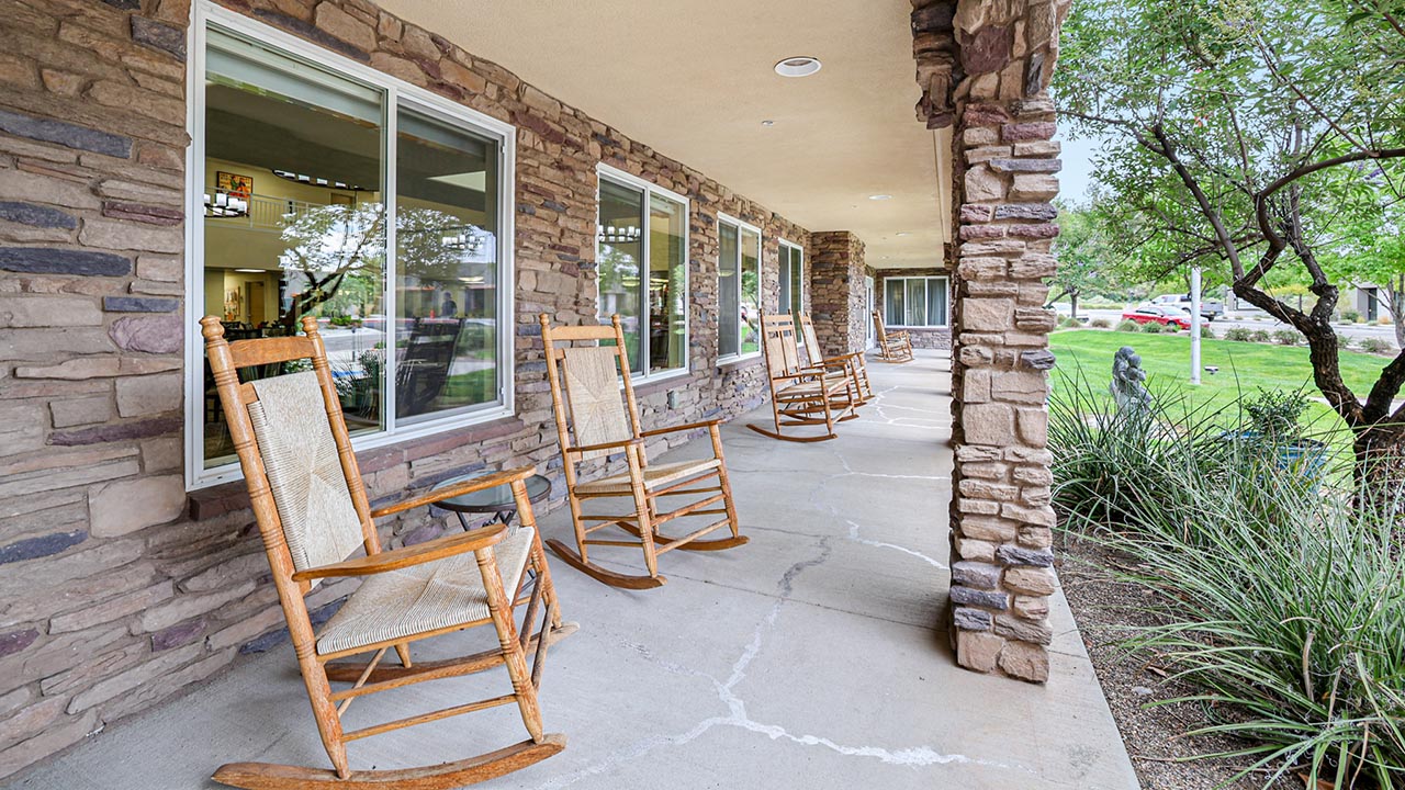 A stone porch with several wooden rocking chairs lined up, large windows reflecting indoor lights, and greenery on the right side, creating a peaceful outdoor seating area.