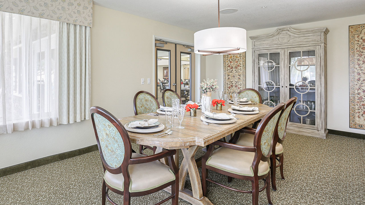 A dining room with a wooden table set for six, elegant upholstered chairs, a modern white pendant light, and a glass-front cabinet against the wall. The room has neutral colors and patterned carpet.