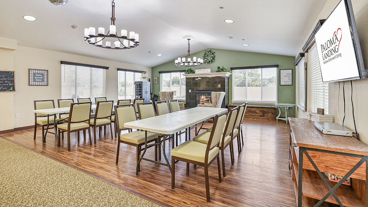 A bright, spacious dining room with several rectangular tables and green chairs, wood flooring, large windows, a fireplace, and a TV displaying the Paloma Landing logo on the right wall.