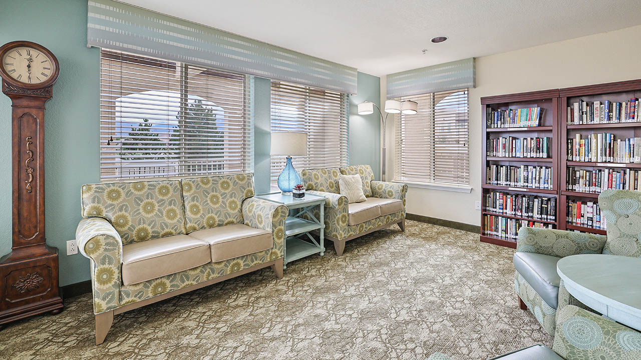 A cozy reading room with two patterned sofas, a blue lamp on a side table, a tall wooden clock, bookshelves filled with books, and large windows with blinds letting in natural light.