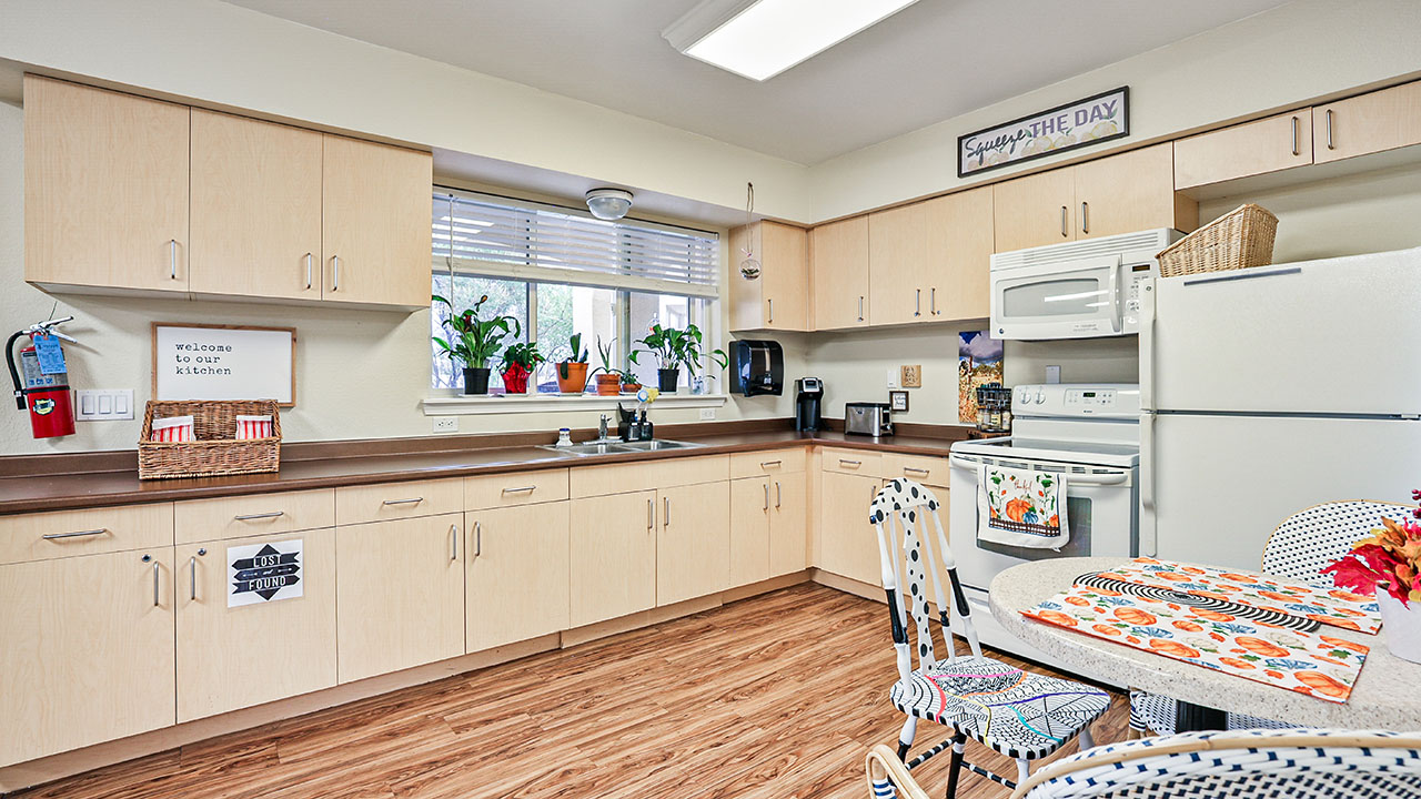 A bright, spacious kitchen with light wood cabinets, white appliances, plants on the windowsill, and a round table with colorful placemats and chairs. A sign reads “Welcome to our kitchen!” and the floor is wood-patterned.