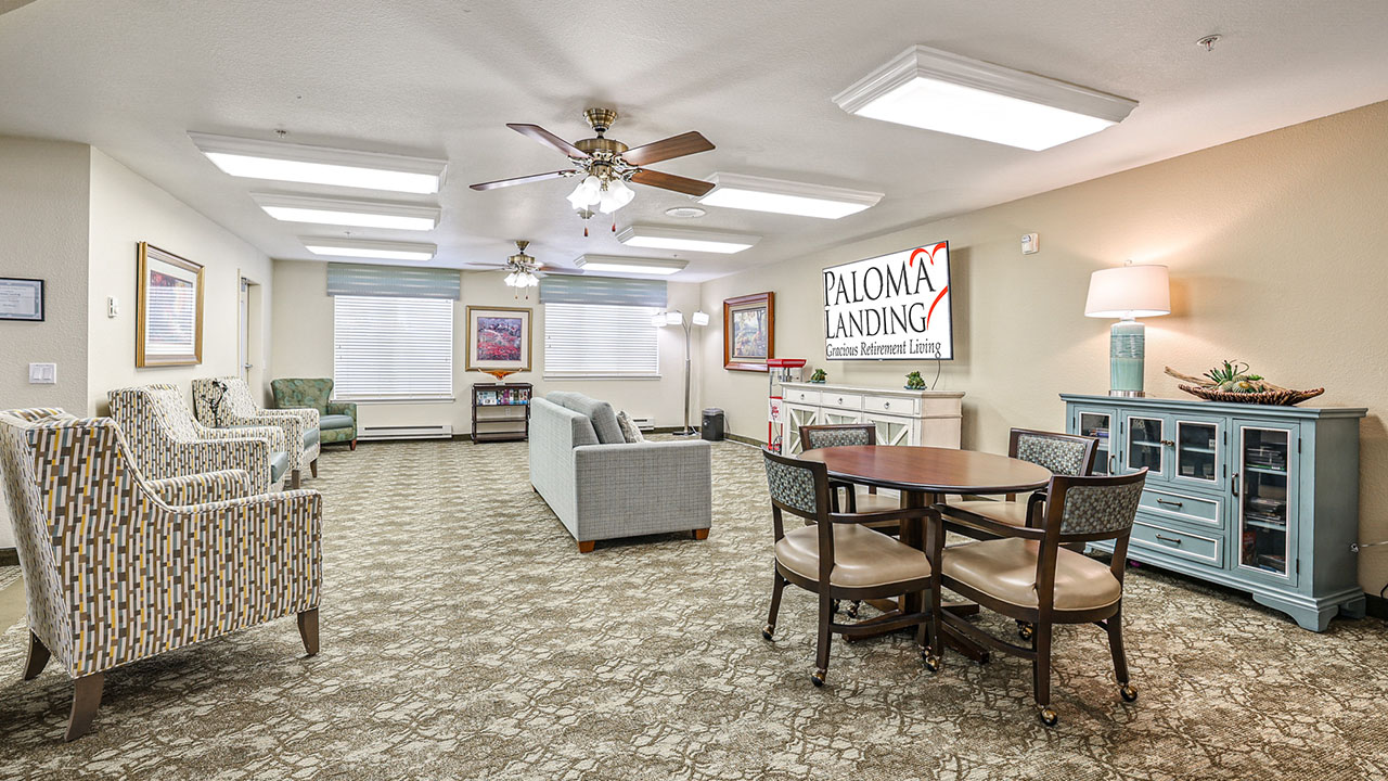 A bright, spacious common area with armchairs, sofas, a round table with chairs, and a blue sideboard. A "Paloma Landing" sign is on the wall, and ceiling fans and lamps provide lighting.