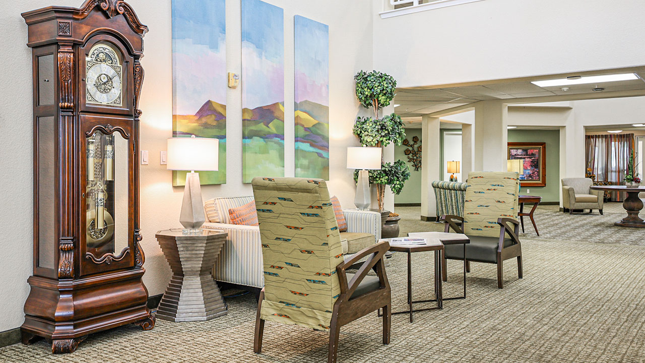A cozy lobby with a large grandfather clock, two patterned armchairs, a small table, and a couch. Abstract mountain artwork and lamps decorate the space, with plants and more seating visible in the background.
