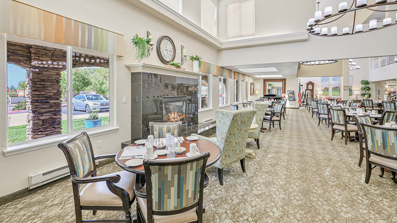 A spacious, well-lit dining area with elegant tables and chairs, set for a meal. Large windows let in natural light, and a fireplace adds warmth. Modern décor and chandeliers create a welcoming atmosphere.