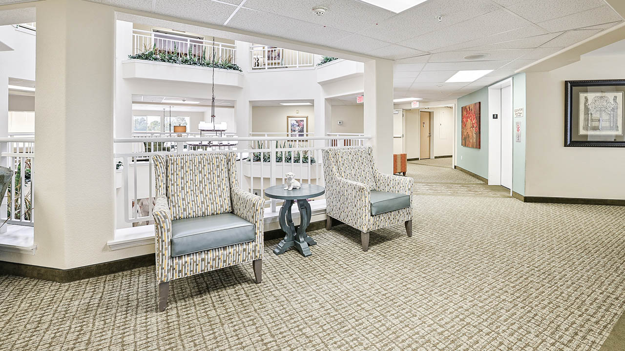Two patterned armchairs and a small round table sit in a bright hallway with light carpet, white railings, and framed artwork on the walls in what appears to be a residential or assisted living facility.
