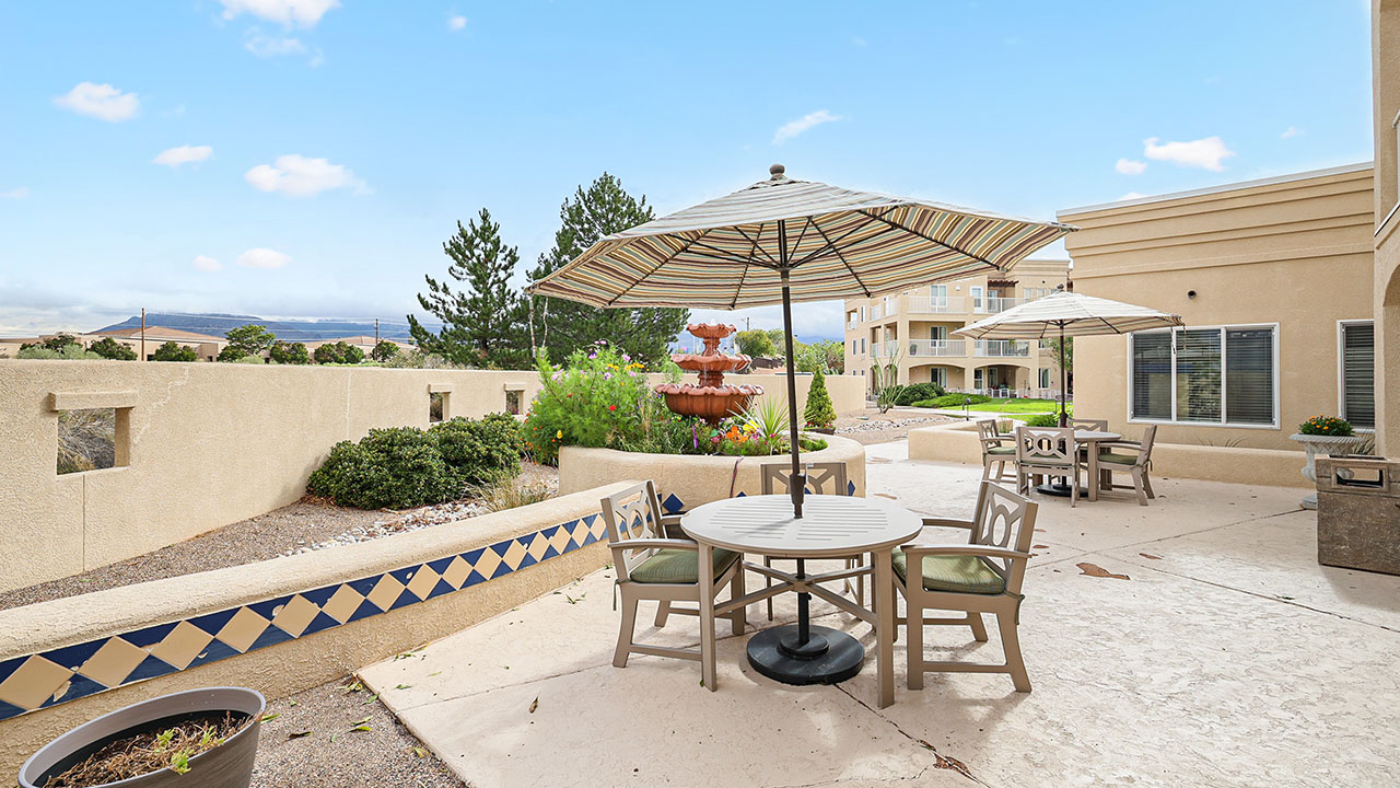 Outdoor patio area with round tables, chairs, and striped umbrellas. A tiered fountain is surrounded by greenery, and beige stucco buildings are in the background under a partly cloudy sky.