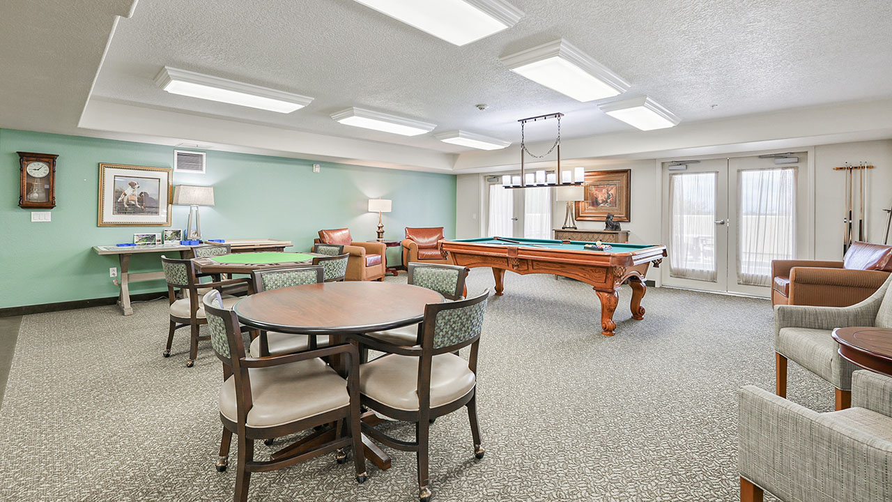 A well-lit recreation room with a billiards table, card table, chairs, armchairs, framed artwork on the walls, and large windows with sheer curtains letting in natural light.