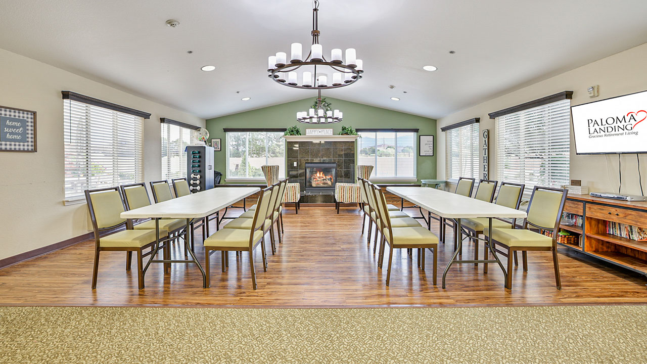 A spacious, well-lit community room with two long tables and chairs arranged in a U-shape, a lit fireplace, large windows, chandeliers, and a TV displaying “Paloma Landing Senior Living.”.