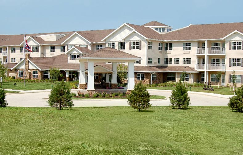 A large, multi-story tan building with a brown roof, featuring a covered entrance supported by white columns. The building is surrounded by a well-maintained lawn and a circular driveway, with a flagpole and trees in the foreground.