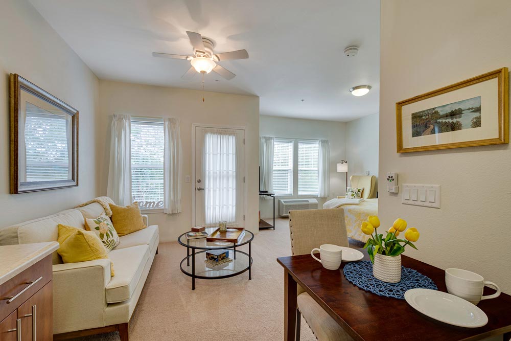 A cozy and brightly lit studio apartment featuring a living area with a white sofa, a glass coffee table, and wall art. A dining table with two cups and a vase of yellow tulips is in the foreground. In the background, a bed is positioned near large windows.