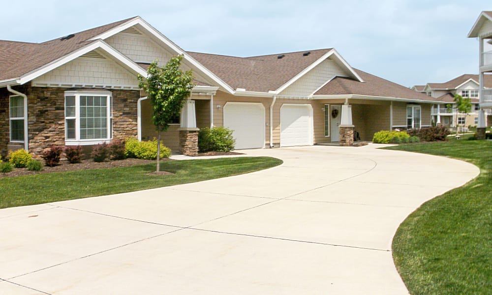 A well-manicured front yard in front of a single-story residential building with brown siding, stone accents, and a tan shingled roof. There are two white garage doors, a wide curved driveway, green grass, small shrubs, and young trees next to the house.