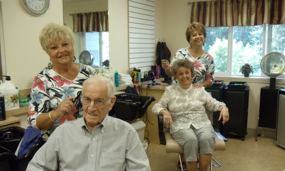 Two elderly individuals are seated in salon chairs, receiving haircuts from two women standing behind them. The salon is well-lit with striped curtains on the windows and various hairdressing tools and products on the counter. Everyone is smiling.