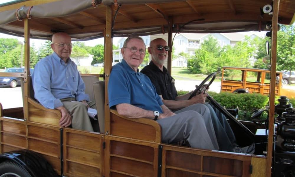 Three elderly men are seated in an old wooden vehicle with open sides. Two are seated in the back while one is behind the wheel. They are all smiling. The background shows a residential area with greenery and parked cars.