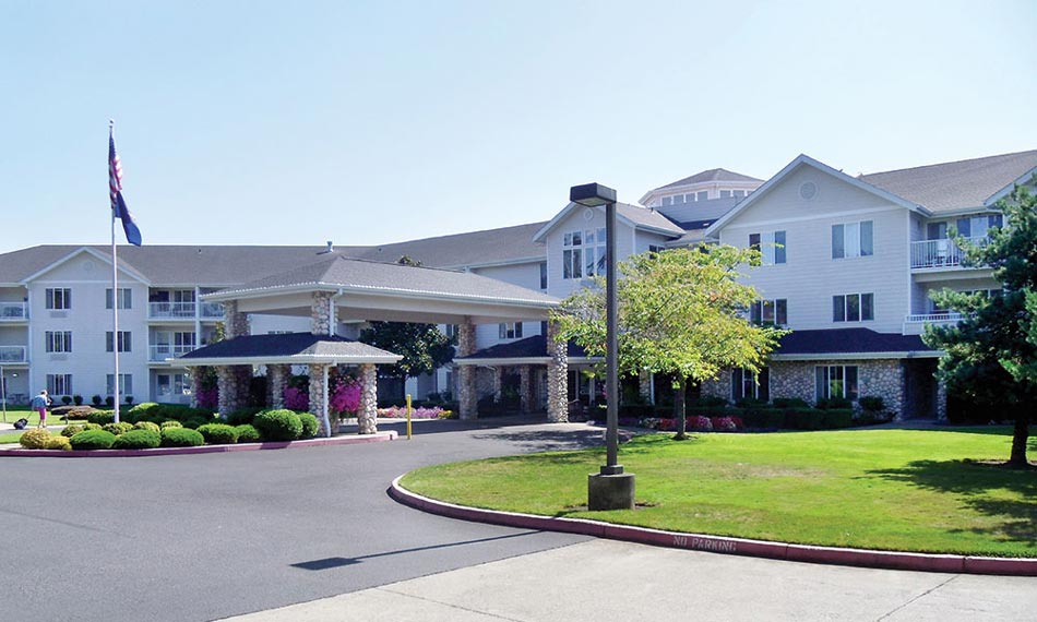 A large, multi-story building with a white exterior and balconies, surrounded by a well-manicured lawn and shrubbery. An American flag flies on a tall pole near the entrance, which has a covered drop-off area. A street lamp and a paved driveway are in the foreground.