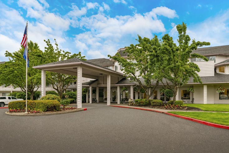 The image shows the exterior of a large, two-story building with white siding and a covered entryway. An American flag stands to the left of the entrance. The surroundings include well-maintained shrubs, trees, and a neatly paved driveway. The sky above is bright and partly cloudy.