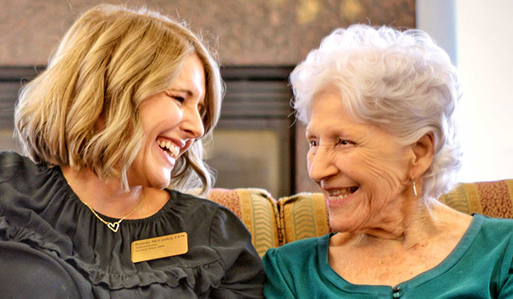 An elderly woman with short white hair and a younger woman with shoulder-length blonde hair share a joyful moment, sitting side by side on a couch, smiling warmly at each other. The younger woman wears a name tag and a dark blouse.