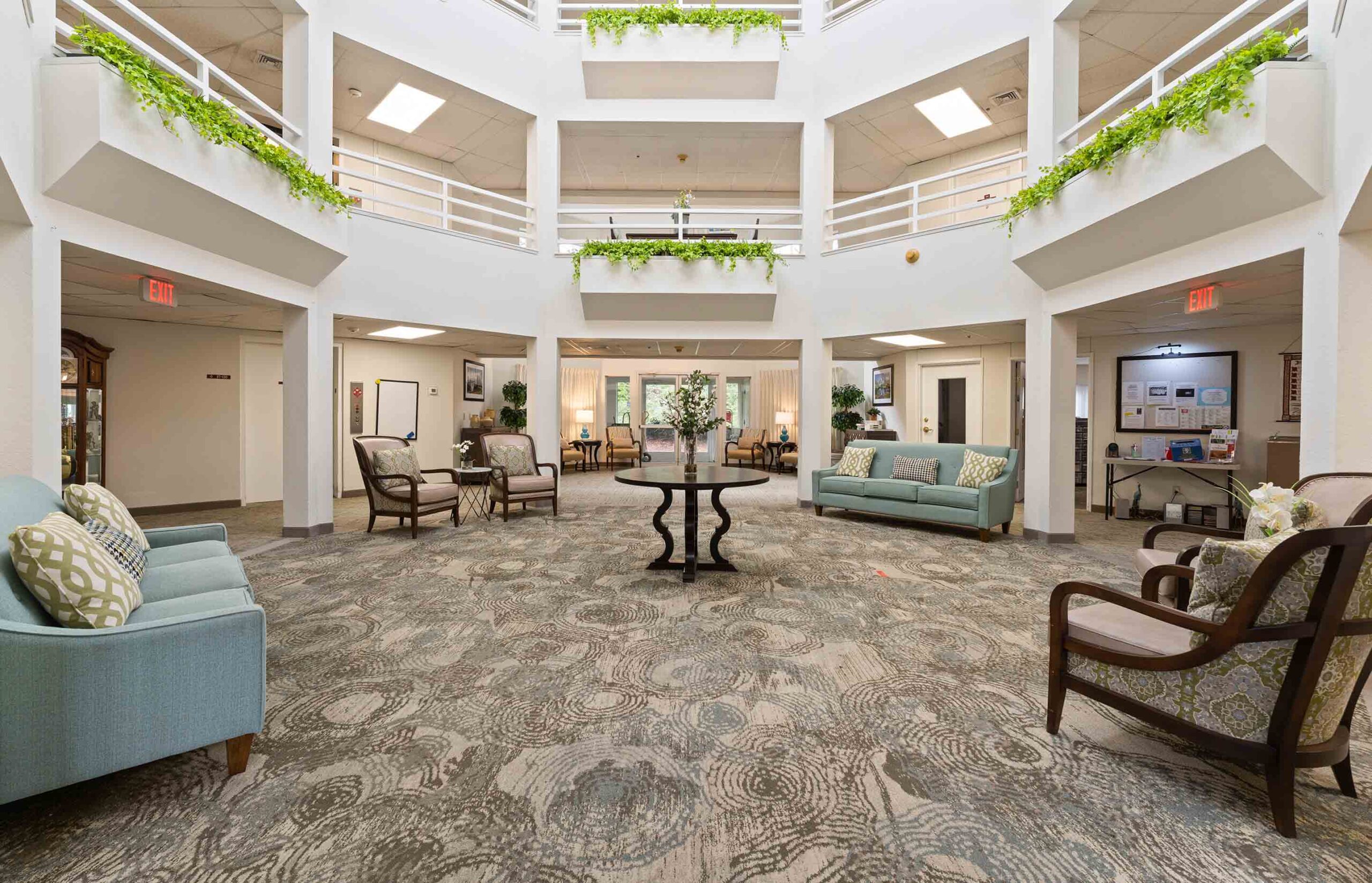 A spacious, well-lit atrium with a high ceiling and multiple levels. The room features comfortable seating with sofas and chairs, a central round table with decorative plants, and potted greenery hanging from the railings of the upper levels.