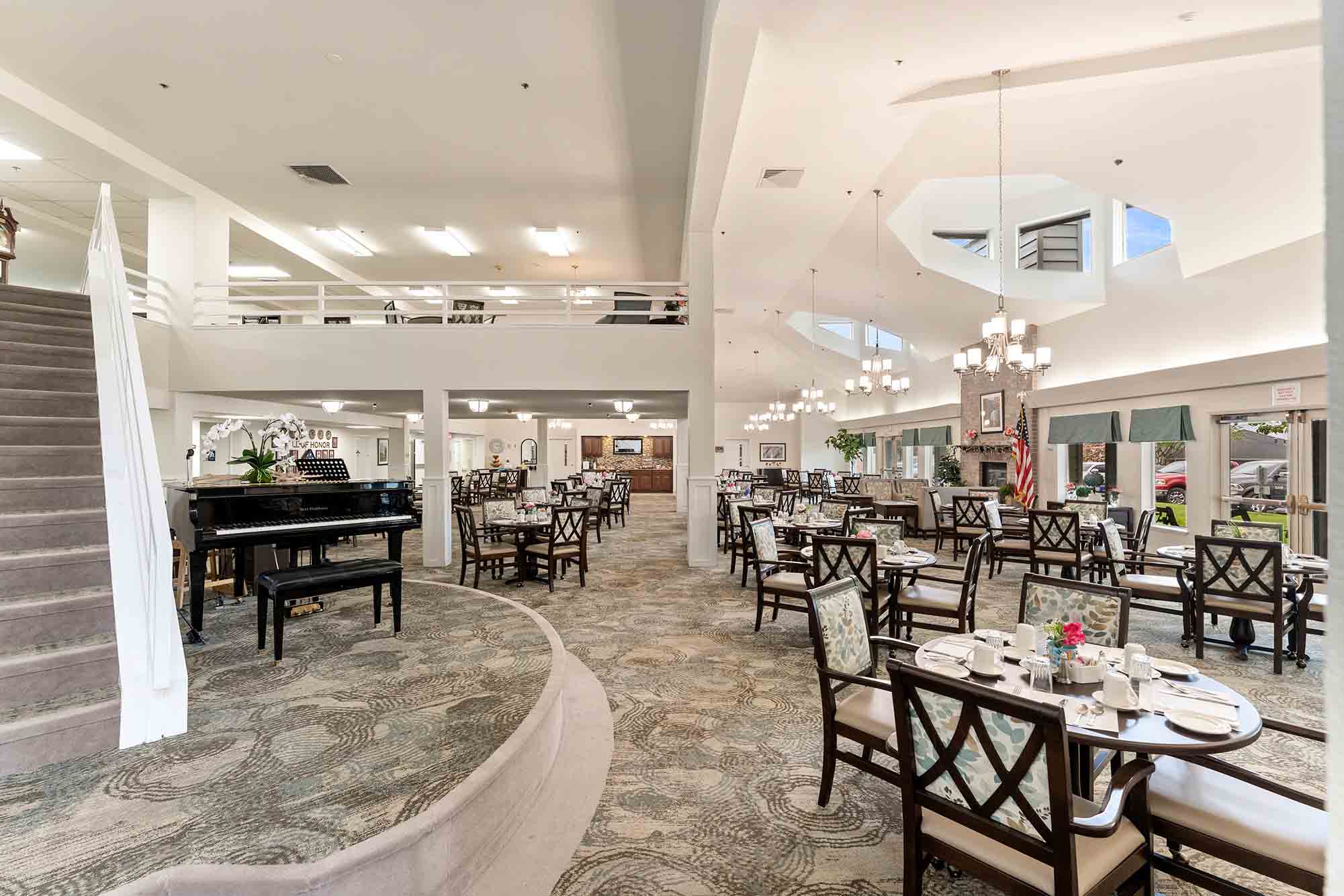 A spacious, elegantly decorated dining area with round and square tables set with white linens and flowers. Chairs are neatly arranged, and a grand piano is positioned near the carpeted staircase leading to an upper level. Chandeliers hang from the high ceiling.