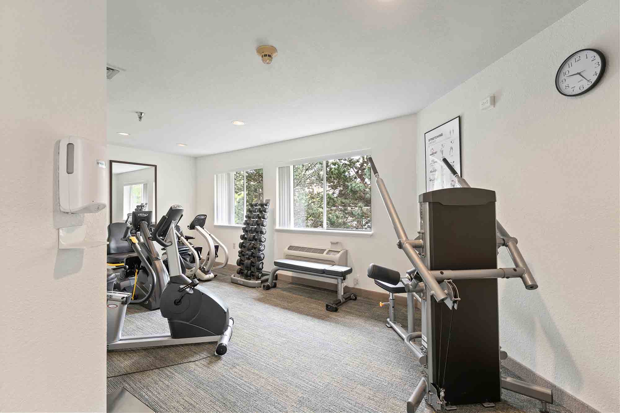 A clean and well-lit gym with exercise equipment including treadmills, stationary bikes, a weight rack, and a multi-function weight machine. The room has large windows letting in natural light, a wall clock, and a hand sanitizer dispenser on the wall.