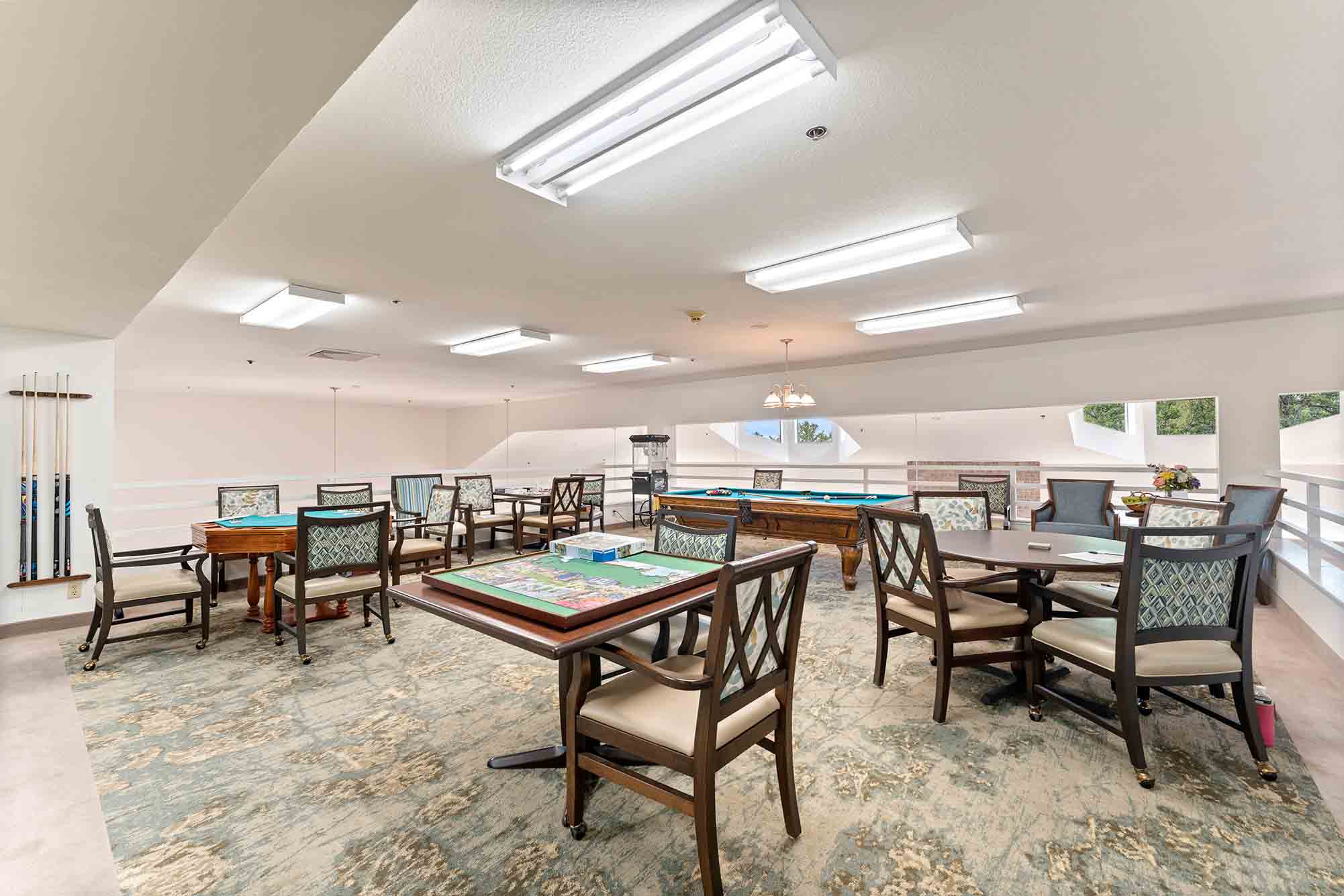 A spacious room with several tables and chairs arranged for board games and card games. A pool table is situated in the far end, and cue sticks are mounted on the wall. The well-lit room has large windows allowing natural light to flood in.