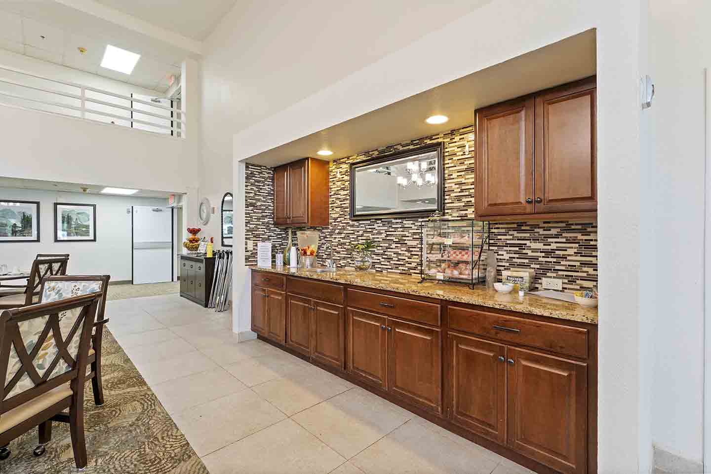 A dining area featuring a long countertop with wooden cabinets below and a tile backsplash. Breakfast items, including a cereal dispenser and fruit, are spread across the counter. There are dining tables and chairs, framed photos on the walls, and a modern chandelier above.