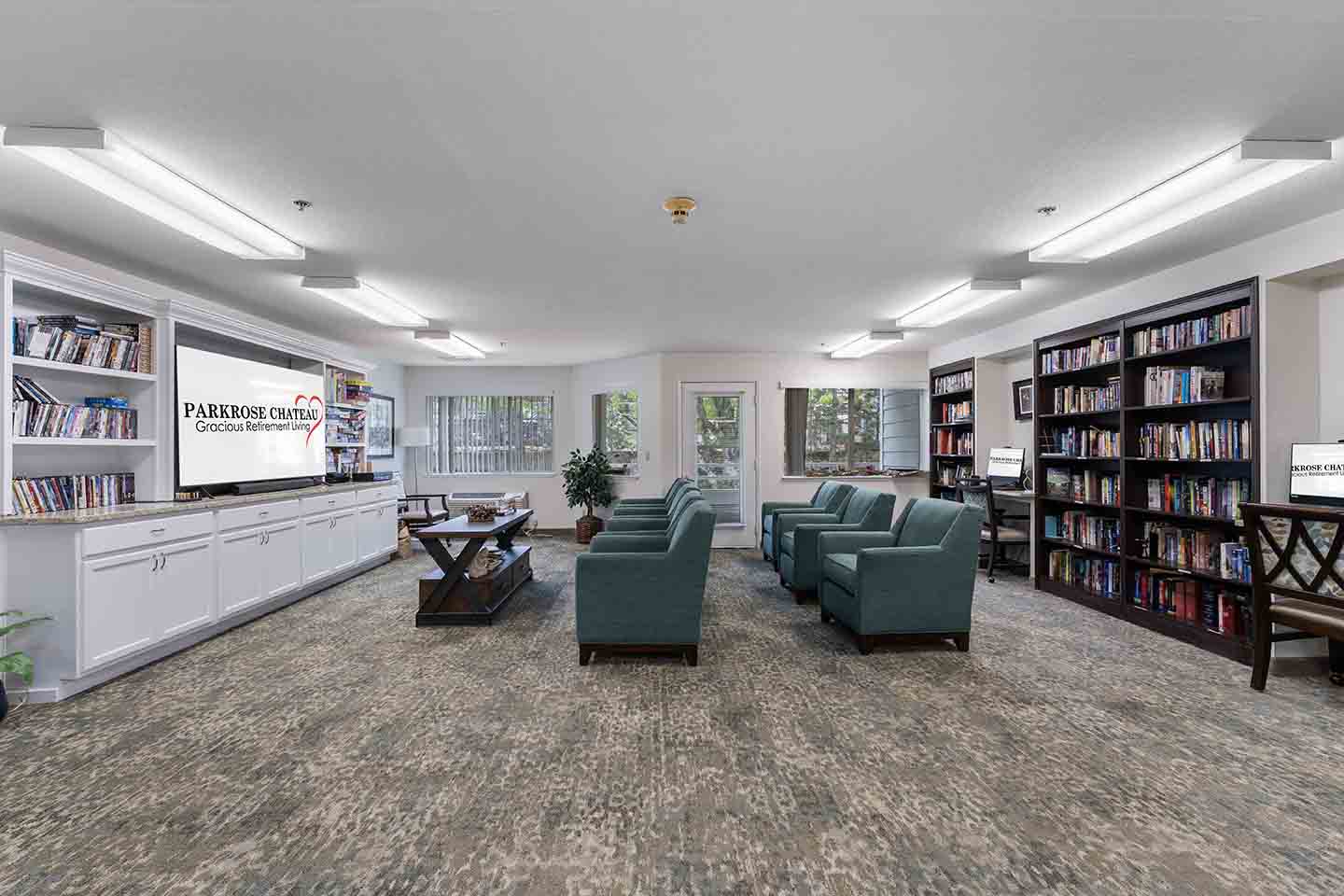 A spacious library with modern lighting features several rows of bookshelves filled with books, a central sitting area with several green armchairs, a large TV on a white cabinet, and multiple windows providing natural light. 