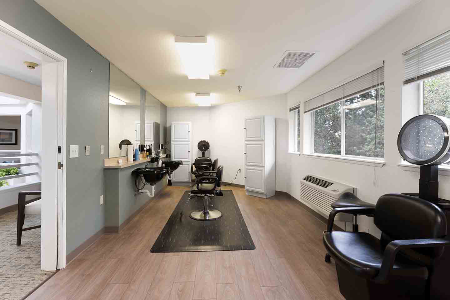 A hair salon with two black styling chairs in front of large mirrors and countertops adorned with various hair products. There's a hairdryer on the wall next to the chairs and a window to the right letting in natural light. The floor is wooden, and a door is open to another room.