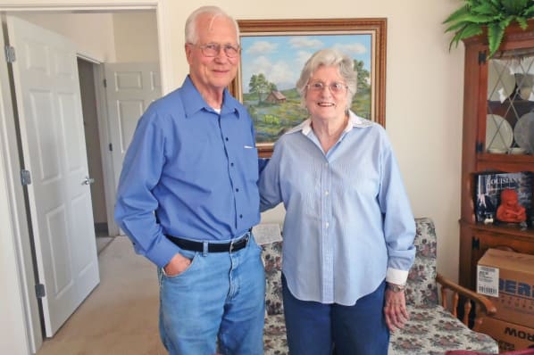 An elderly couple smiles while standing in a living room. The man wears glasses, a blue shirt, and jeans, with one hand in his pocket. The woman wears glasses and a light blue blouse. A painting, bookshelf, and an open door are visible in the background.