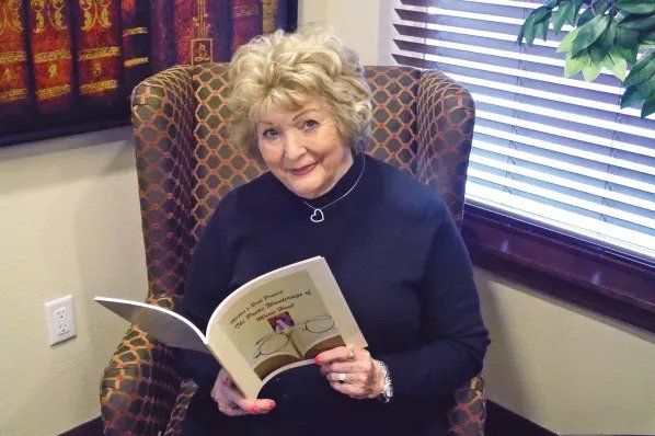 An elderly woman with short, curly blonde hair is sitting in a patterned armchair, holding an open book and smiling at the camera. She is wearing a black top and a necklace with a heart pendant. A window with blinds and a framed decoration are in the background.