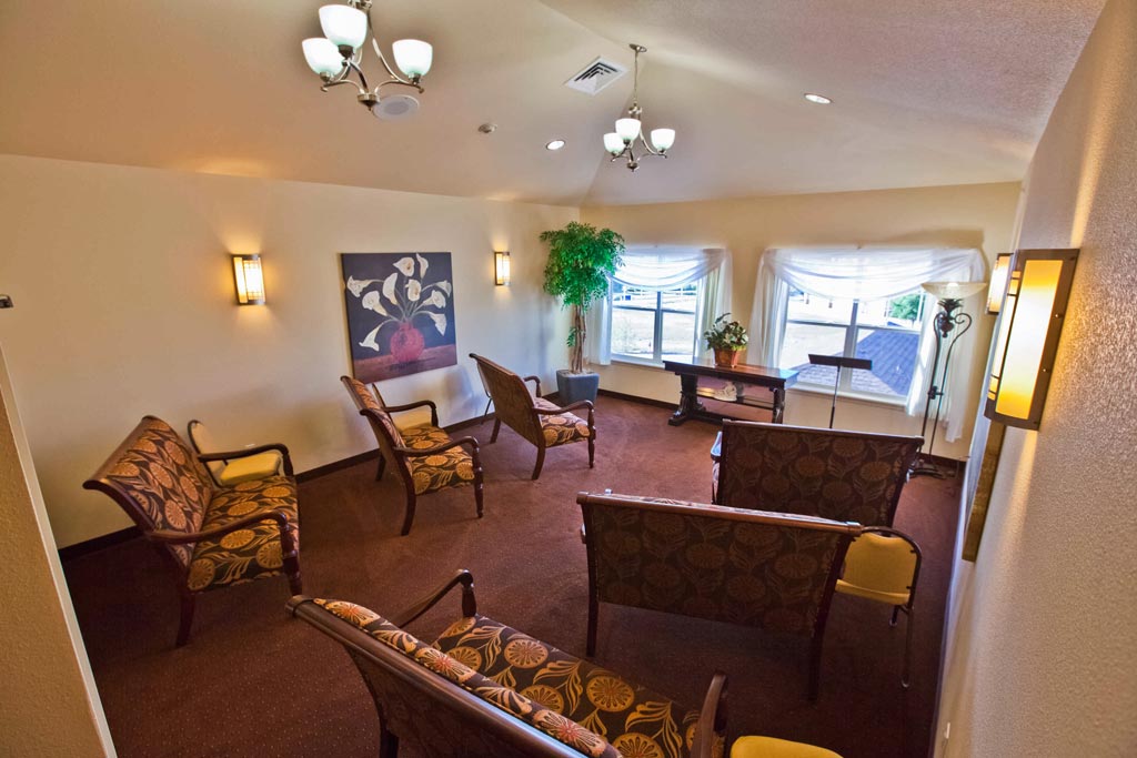 A small room with several cushioned chairs arranged in rows facing a lectern, possibly for a meeting or small gathering. The walls are adorned with framed artwork and two windows with sheer white curtains. The room is softly lit by overhead lights and wall sconces.