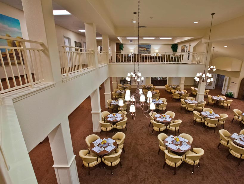 A spacious dining area with beige walls and carpet, featuring round tables covered with plates and cups, each surrounded by four cream-colored chairs. Elegant chandeliers hang from the high ceiling, and a second-floor balcony overlooks the room.