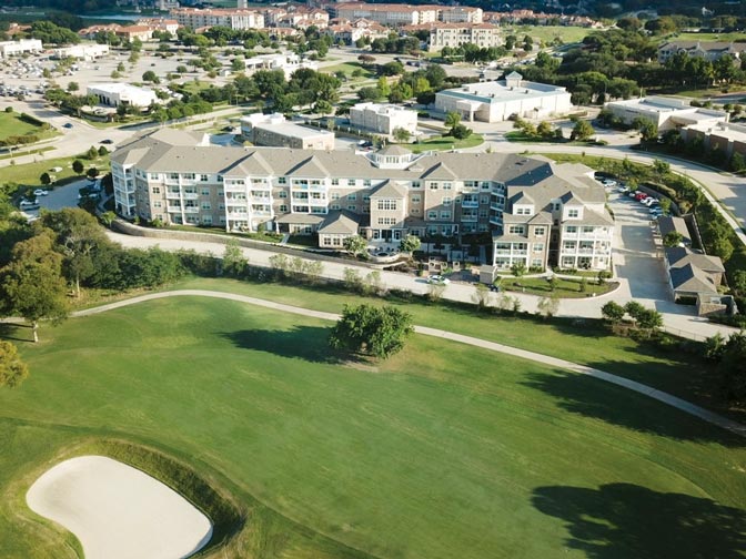 Aerial view of a residential complex surrounded by grassy lawns, trees, and adjacent to a golf course with a sand bunker. Multiple buildings with beige exteriors and parking lots are visible, with a background of more buildings and greenery in the distance.