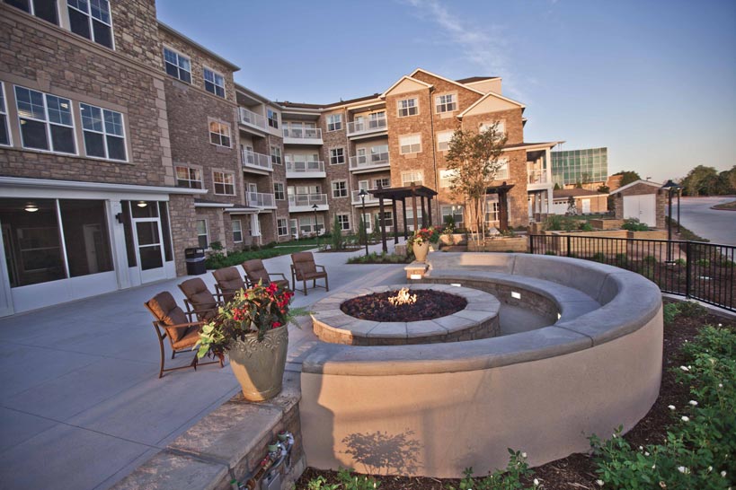 Outdoor patio area with circular fire pit surrounded by chairs. There is a large modern apartment building with balconies in the background. The patio features potted plants, lush landscaping, and clear skies above.