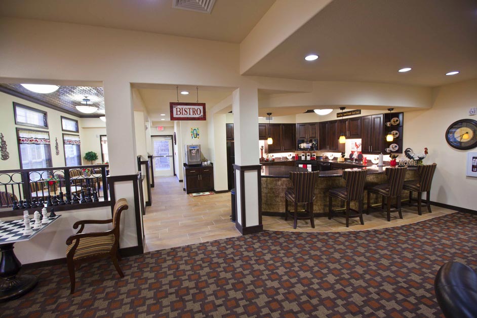 A cozy bistro-style kitchen and dining area with warm lighting, dark wood cabinets, and a large counter with bar stools. Adjacent seating areas feature a chessboard table and elegant chairs. The space is decorated with inviting accents and wall art.