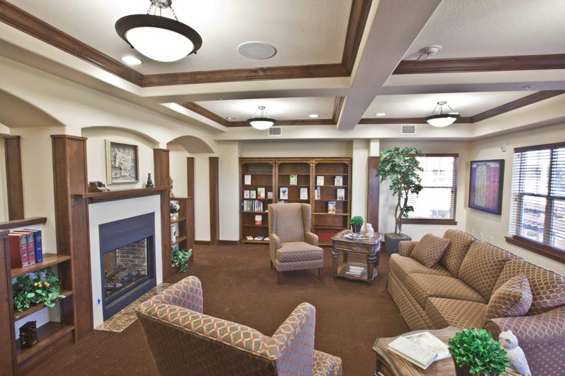 A cozy living room with brown carpet, patterned armchairs, and sofas. The room features a fireplace with built-in shelves on the sides, filled with books and decor. There's a large potted plant in the corner and ample natural light from the windows.