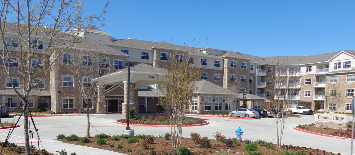 A large, multi-story residential building with a well-maintained landscape, including young trees, bushes, and flowers. A driveway with parked cars leads to the main entrance. A sign reads 