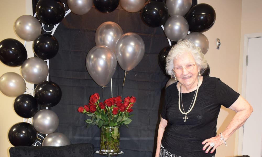 An elderly woman wearing a black outfit with pearl jewelry poses in front of a black and silver balloon arch. Next to her is a vase of red roses. She is smiling and standing with her hand on her hip. The background is indoor with a neutral-colored wall and a door.